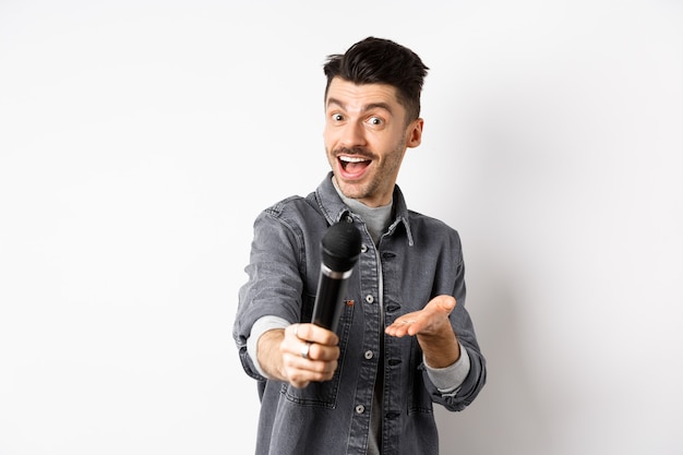 Homem bonito e sorridente estende a mão com o microfone, dando-lhe o microfone, de pé sobre um fundo branco.