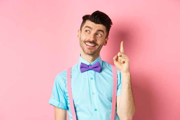 Homem bonito e sorridente, levantando o dedo em gesto de eureka, desviando o olhar e lançando uma ideia, tem uma sugestão ou um plano, de pé na gravata borboleta sobre fundo rosa.