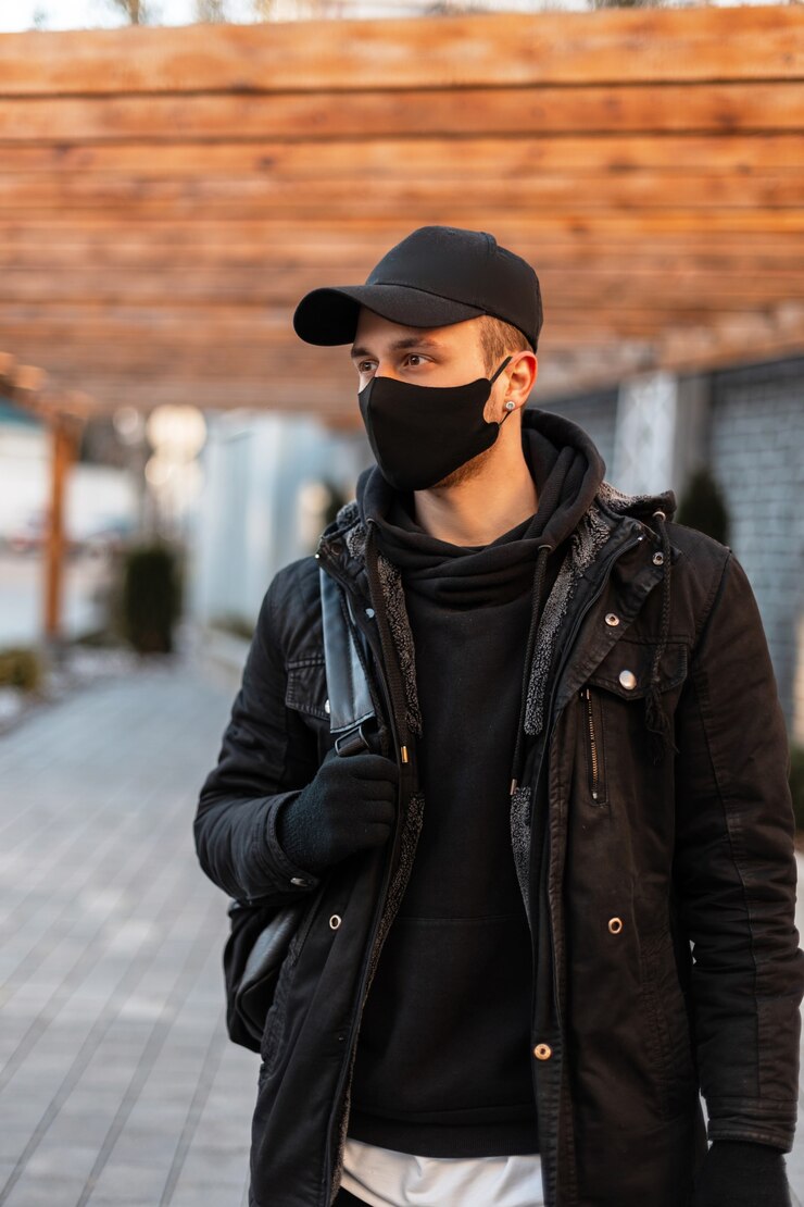 Foto homem bonito elegante com uma máscara protetora em roupas da moda com uma jaqueta, capuz, boné de maquete e mochila caminha na cidade. estilo de roupa urbana masculina