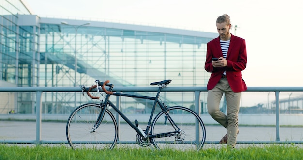 Homem bonito elegante jovem caucasiano em pé na rua com a bicicleta. olhando para o relógio de mão e mensagens de texto no smartphone. macho tocando e rolando no celular com bicicleta lá fora. esperando.