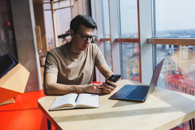 Homem bonito em camiseta casual vermelha, escrevendo uma mensagem no celular enquanto está sentado perto da janela de vidro na mesa com o laptop trabalhando remotamente no café