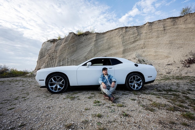 Homem bonito em jaqueta jeans e boné sentado perto de seu muscle car branco na carreira