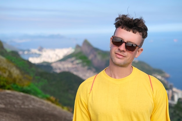 Homem bonito em óculos de sol sorri no fundo das montanhas e oceano