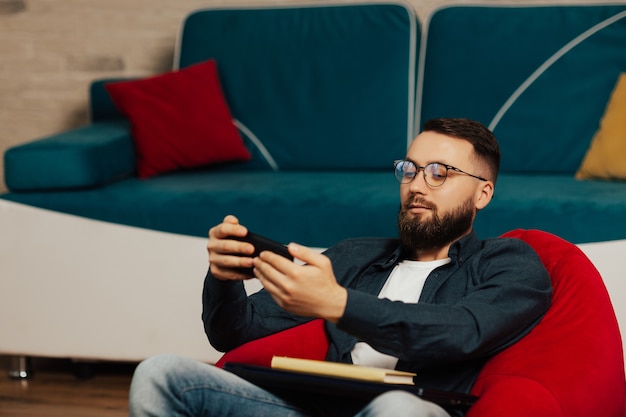 Homem bonito está usando o dispositivo smartphone enquanto está sentado na poltrona em casa.