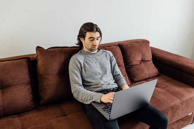 Homem bonito estudante caucasiano sentado no sofá e aprendendo com o laptop e livros, cansado