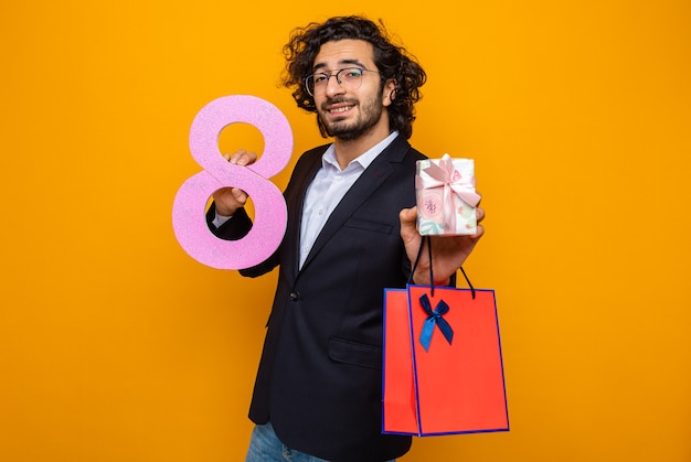 Homem bonito feliz e positivo de terno segurando o presente saco de papel com o presente e o número oito sorrindo confiante celebrando o dia internacional da mulher, 8 de março