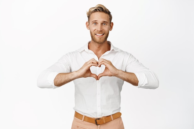 Homem bonito feliz mostrando que eu te amo sinal de gesto de coração e grande sorriso alegre em pé sobre fundo branco