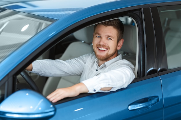 Homem bonito feliz sorrindo alegremente para a câmera enquanto está sentado em seu novo carro na concessionária