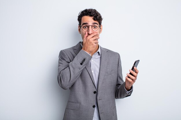 Homem bonito hispânico cobrindo a boca com as mãos com um choque. conceito de negócios e telefone