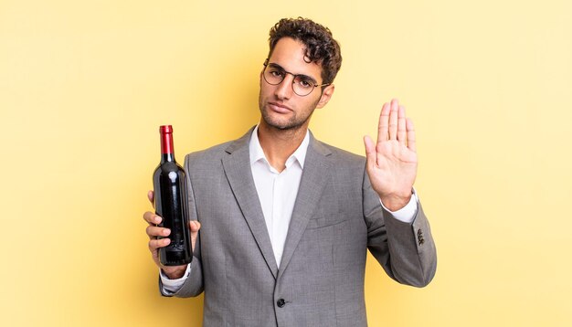 Homem bonito hispânico olhando sério mostrando a palma da mão aberta, fazendo gesto de parada. conceito de garrafa de vinho