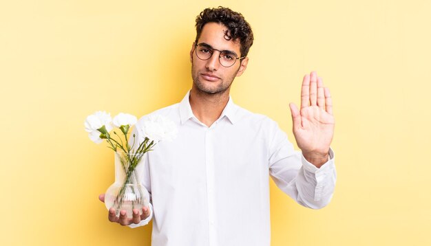 Homem bonito hispânico olhando sério mostrando a palma da mão aberta, fazendo gesto de parada. conceito de vaso de flores