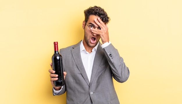 Homem bonito hispânico parecendo chocado, assustado ou apavorado, cobrindo o rosto com a mão. conceito de garrafa de vinho