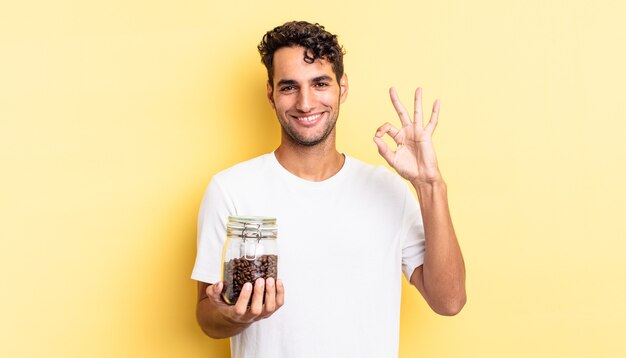 Homem bonito hispânico se sentindo feliz, mostrando aprovação com um gesto certo. garrafa de grãos de café