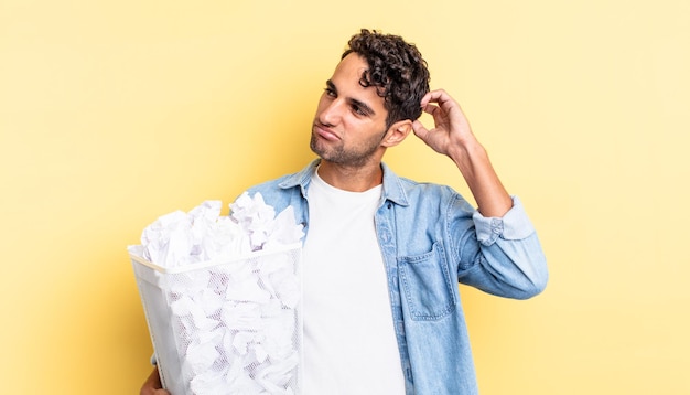 Homem bonito hispânico se sentindo perplexo e confuso, coçando a cabeça. conceito de lixo de bolas de papel