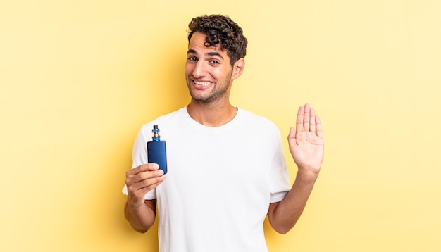 Homem bonito hispânico sorrindo alegremente, acenando com a mão, dando as boas-vindas e cumprimentando você. conceito de vaporizador