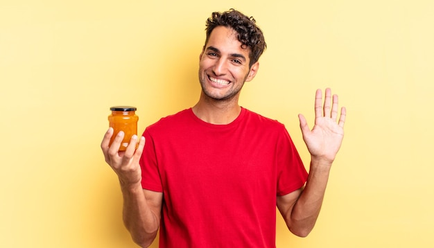 Homem bonito hispânico sorrindo alegremente, acenando com a mão, dando as boas-vindas e cumprimentando você. geléia de pêssego