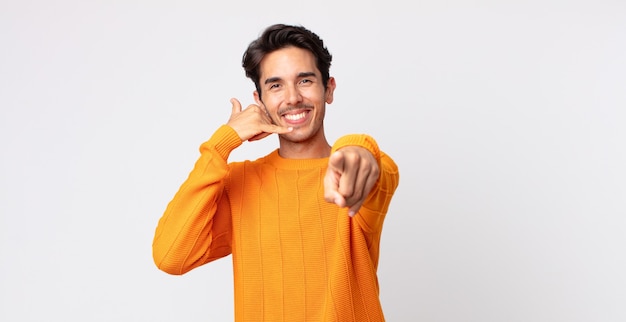 Homem bonito hispânico sorrindo alegremente e apontando para a câmera enquanto faz um gesto para ligar para você, falando ao telefone