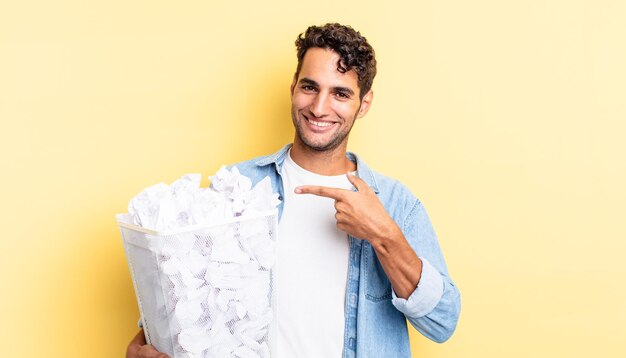 Homem bonito hispânico sorrindo alegremente, sentindo-se feliz e apontando para o lado. conceito de lixo de bolas de papel