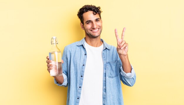 homem bonito hispânico sorrindo e parecendo feliz, gesticulando vitória ou paz. conceito de garrafa de água