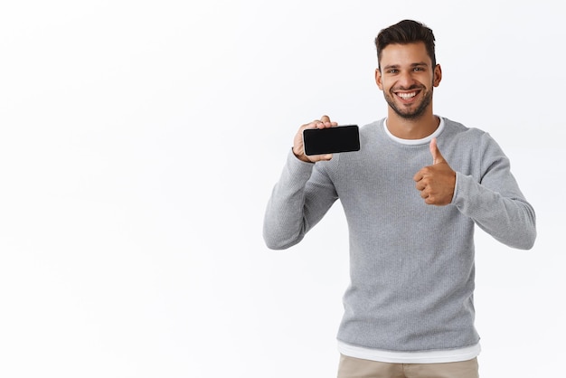 Homem bonito impressionado e satisfeito com cerdas no suéter cinza mostra o gesto de polegar como recomendar o aplicativo para smartphone mostrando a tela do celular horizontalmente satisfeito com o novo fundo branco do jogo