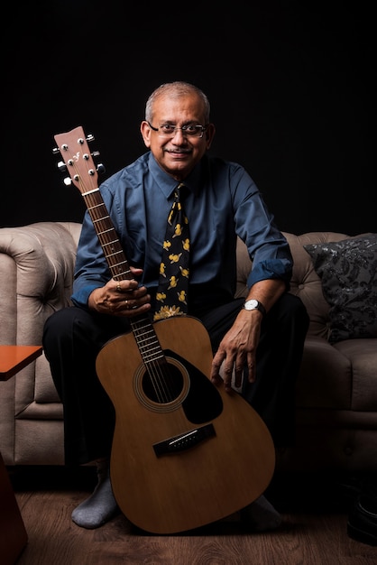 Homem bonito indiano asiático sênior tocando ou posando com um instrumento musical de cordas, como violão ou violino, em pé sobre um fundo preto ou sentado no sofá ou sofá