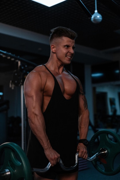 Foto homem bonito jovem atleta forte emocional com um corpo musculoso e uma camiseta preta faz um exercício e treino no ginásio em um fundo escuro. estilo de vida saudável