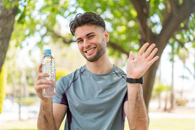 Homem bonito jovem esporte com uma garrafa de água ao ar livre saudando com a mão com expressão feliz