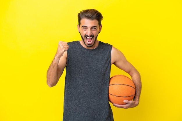 Homem bonito jovem jogador de basquete isolado em fundo amarelo comemorando uma vitória na posição de vencedor