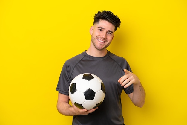 Homem bonito jovem jogador de futebol isolado em um fundo amarelo surpreso e apontando para a frente