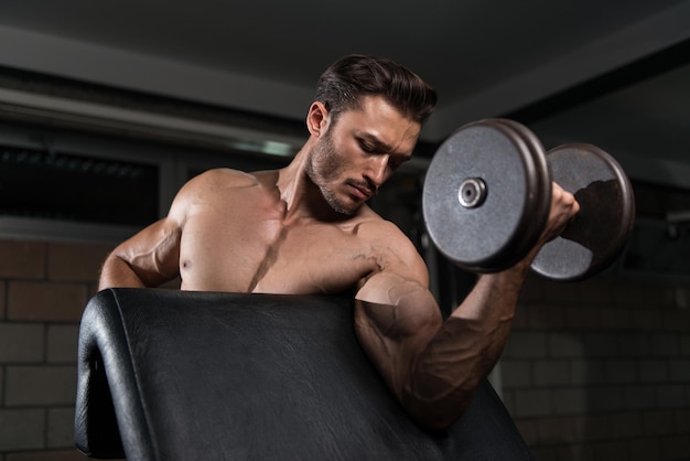 Homem bonito malhando bíceps em um ginásio escuro Dumbbell concentração cachos