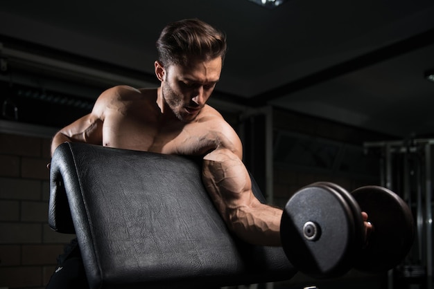 Homem bonito malhando bíceps em um ginásio escuro Dumbbell concentração cachos