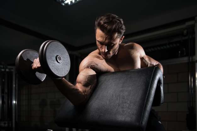 Homem bonito malhando bíceps em um ginásio escuro Dumbbell concentração cachos