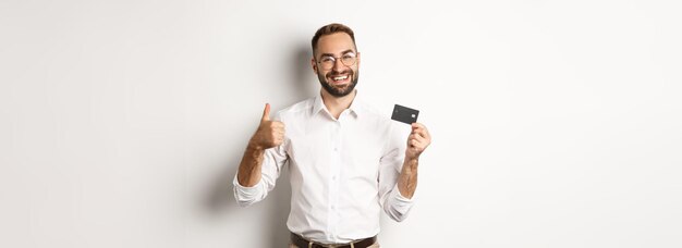 Homem bonito, mostrando seu cartão de crédito e polegar para cima, recomendando banco em pé sobre fundo branco