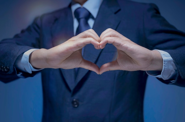 Homem bonito mostrar o símbolo do coração, ícone do amor para amante de negócios, eu amo meu trabalho, cuidar do conceito de serviço. Homem de negócios inteligente usa camisa de terno azul e gravata fazendo formato de coração com a mão sobre fundo azul