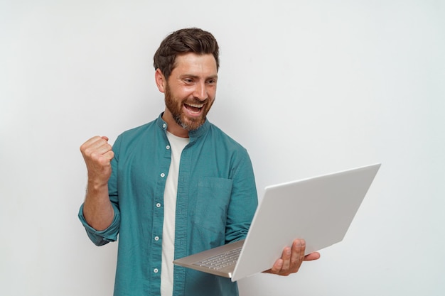 Foto homem bonito muito feliz olha para laptop com punho levantado triunfo no fundo branco