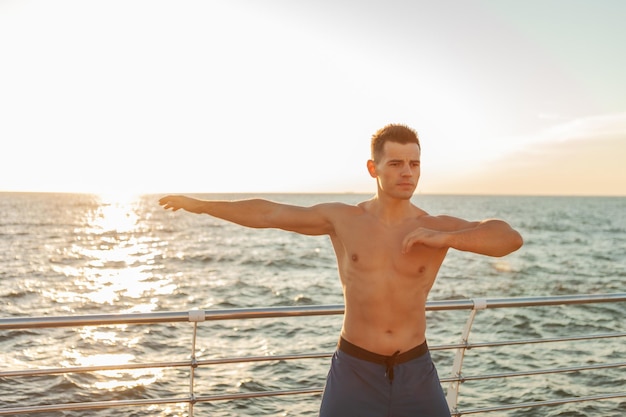 Homem bonito musculoso com torso nu fazendo exercício de aquecimento antes do treino na praia ao nascer do sol