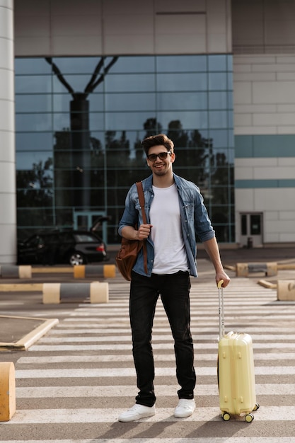 Homem bonito na camiseta branca de jaqueta jeans e calça preta sorri Turista em óculos de sol segura mala amarela e mochila marrom