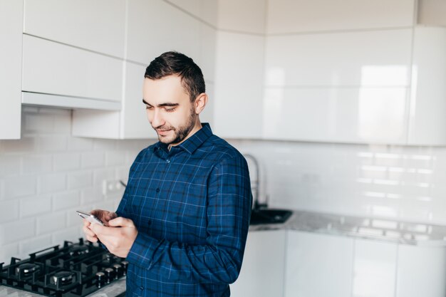 Homem bonito na cozinha sorrindo pela manhã e olhando para o telefone inteligente