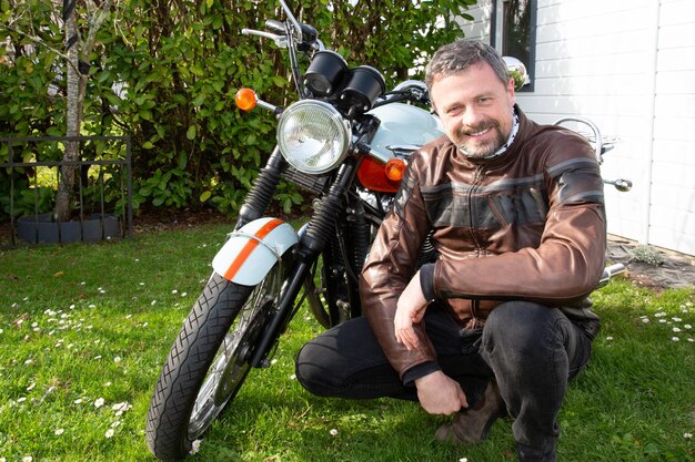 Homem bonito na jaqueta de moda de moto de couro marrom ao lado de motocicleta retrô vintage ao ar livre