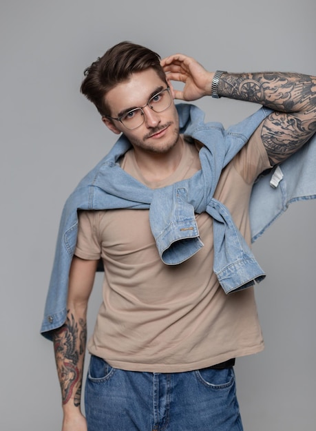 Homem bonito na moda com óculos e cabelo em roupas da moda com uma camiseta e camisa jeans com tatuagens nos braços no estúdio