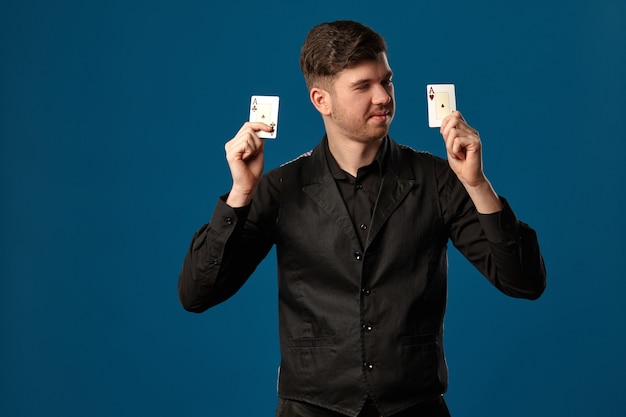 Homem bonito, novato no pôquer, de colete e camisa pretos. Segurando duas cartas de jogar, sorrindo e olhando para elas. Posando contra o fundo azul do estúdio. Jogos de azar, cassino. Fechar-se.