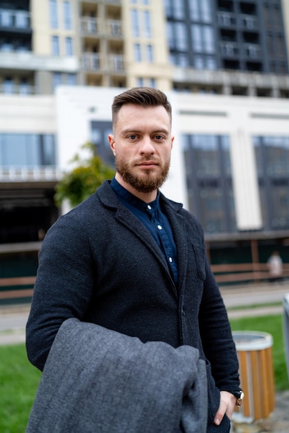 Homem bonito olha para a câmera enquanto usava um terno formal e segurando o casaco cinza nas mãos. Edifício moderno ao fundo.