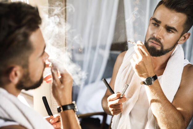 Homem bonito olhando no espelho, fumando um cigarro e raspando a barba com uma navalha