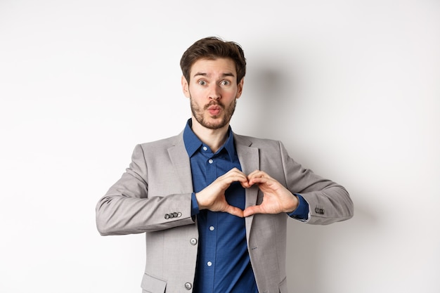 Homem bonito parecendo bobo, mostrando sinal de amor de coração e lábios franzidos, esperando o beijo do amante, de terno em fundo branco.