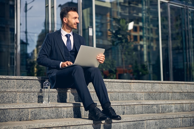 Homem bonito passando um tempo fora do centro de negócios nas escadas enquanto trabalhava on-line no laptop