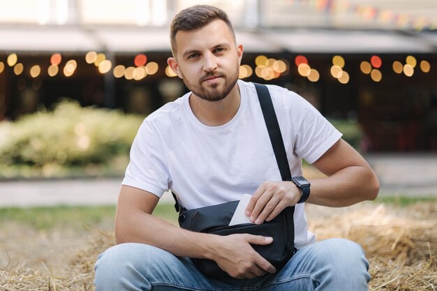 Homem bonito pega o telefone da bolsa tiracolo Homem sentado em um banco de feno ao ar livre