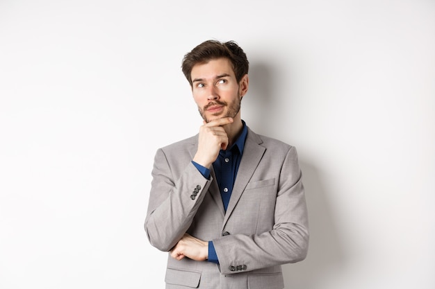 Homem bonito pensativo de terno olhando para cima e pensando, fazendo escolhas, ponderando ideias, de pé contra um fundo branco.