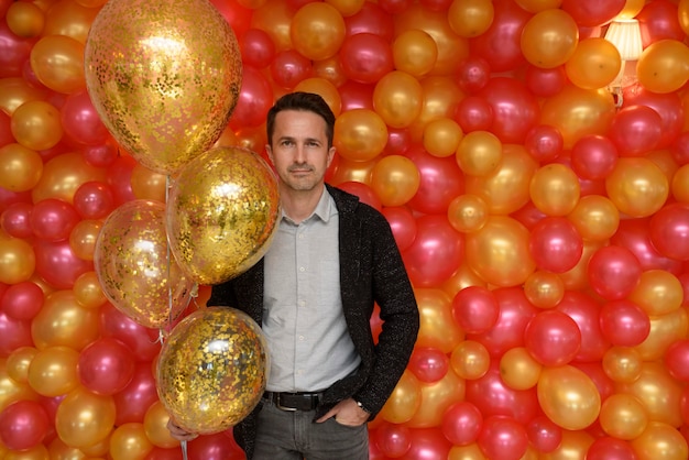 Homem bonito posando com bolas douradas na parede do fundo de fotos de balões vermelhos e amarelos