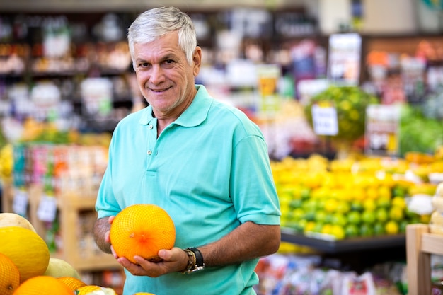 Homem bonito posando, olhando para a câmera e segurando o melão na mão. Cliente barbudo sorrindo. Seção com cítricos frescos no espaço.