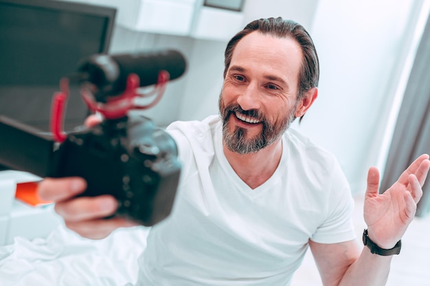 Homem bonito positivo sorrindo e acenando com a mão enquanto segura uma câmera com um microfone no topo e grava a si mesmo